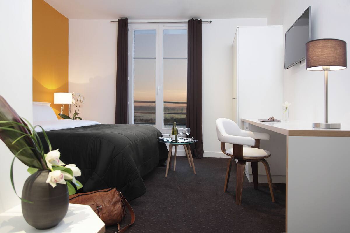 A hotel room with a bed, a desk, a chair, a small table, a vase with flowers, a lamp, and a TV.