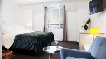 A hotel room with a bed, a chair, a small table, a TV, and a window with curtains.