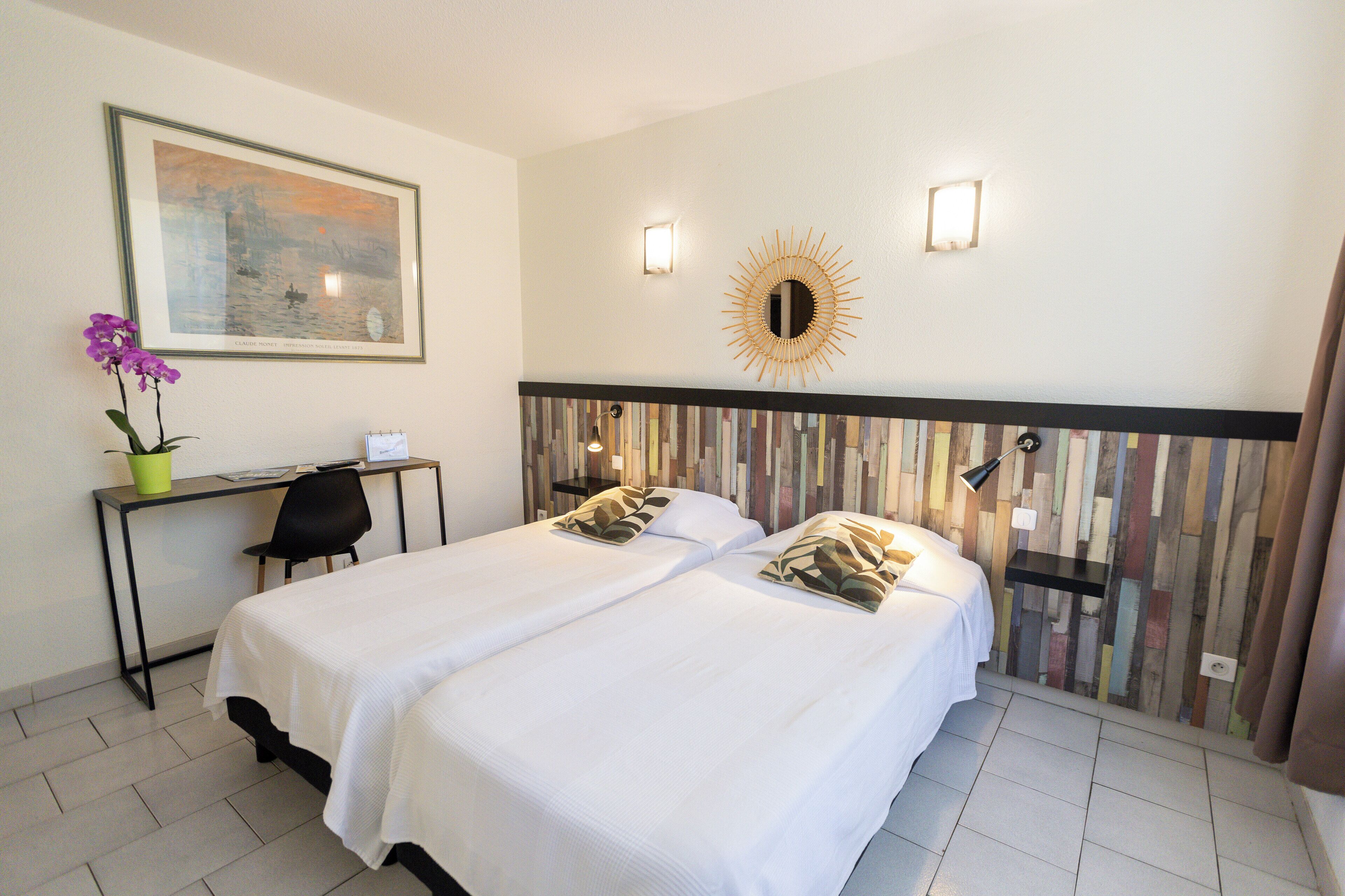 A hotel room with two beds, a desk, a chair, a potted plant, and a painting on the wall.
