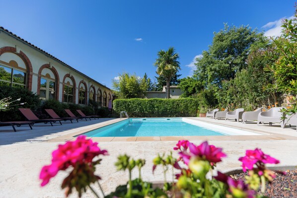 Una piscina con sillas de descanso, un edificio con ventanales arqueados y abundante vegetación.