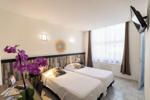 Habitación de hotel con dos camas, un escritorio y una silla, una pequeña mesa con un jarrón de flores, un espejo en la pared y una ventana con cortinas.