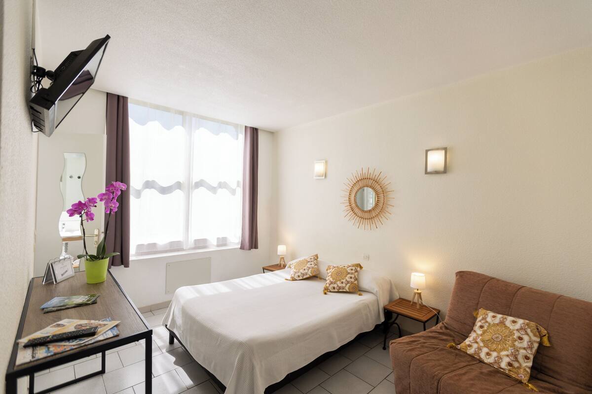 Una habitación de hotel con una cama, un escritorio con lámpara, un televisor, una ventana con cortinas y un espejo en la pared.