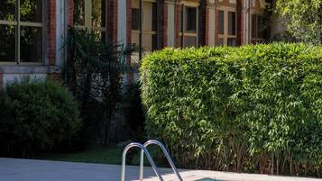 Ein Swimmingpool mit Metallleiter, umgeben von einer Hecke und einem Gebäude mit Rundbogenfenstern und einem Ziegeldach.