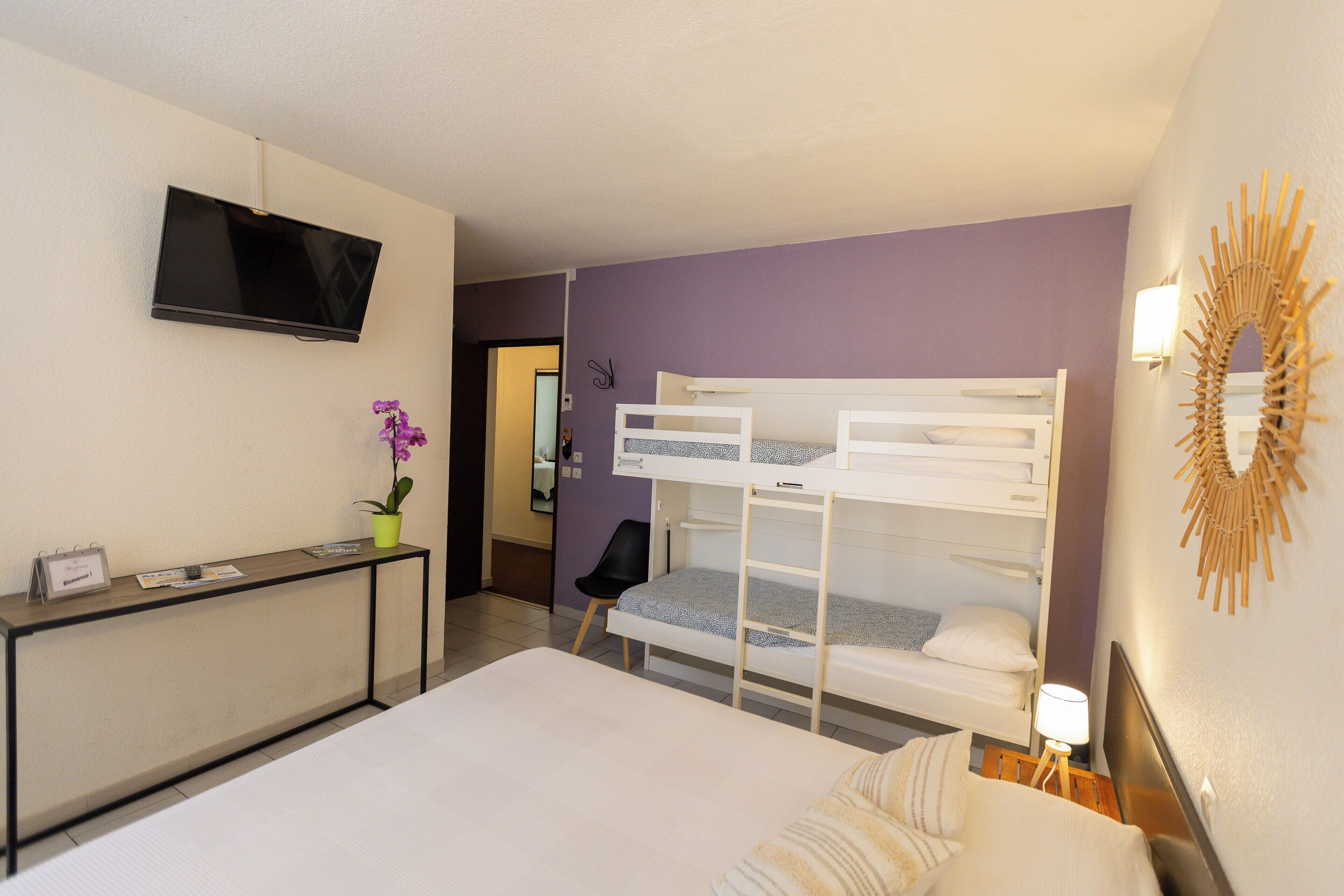 A hotel room with a bunk bed, a desk with a flat-screen TV, a small table with a lamp, and a potted plant.