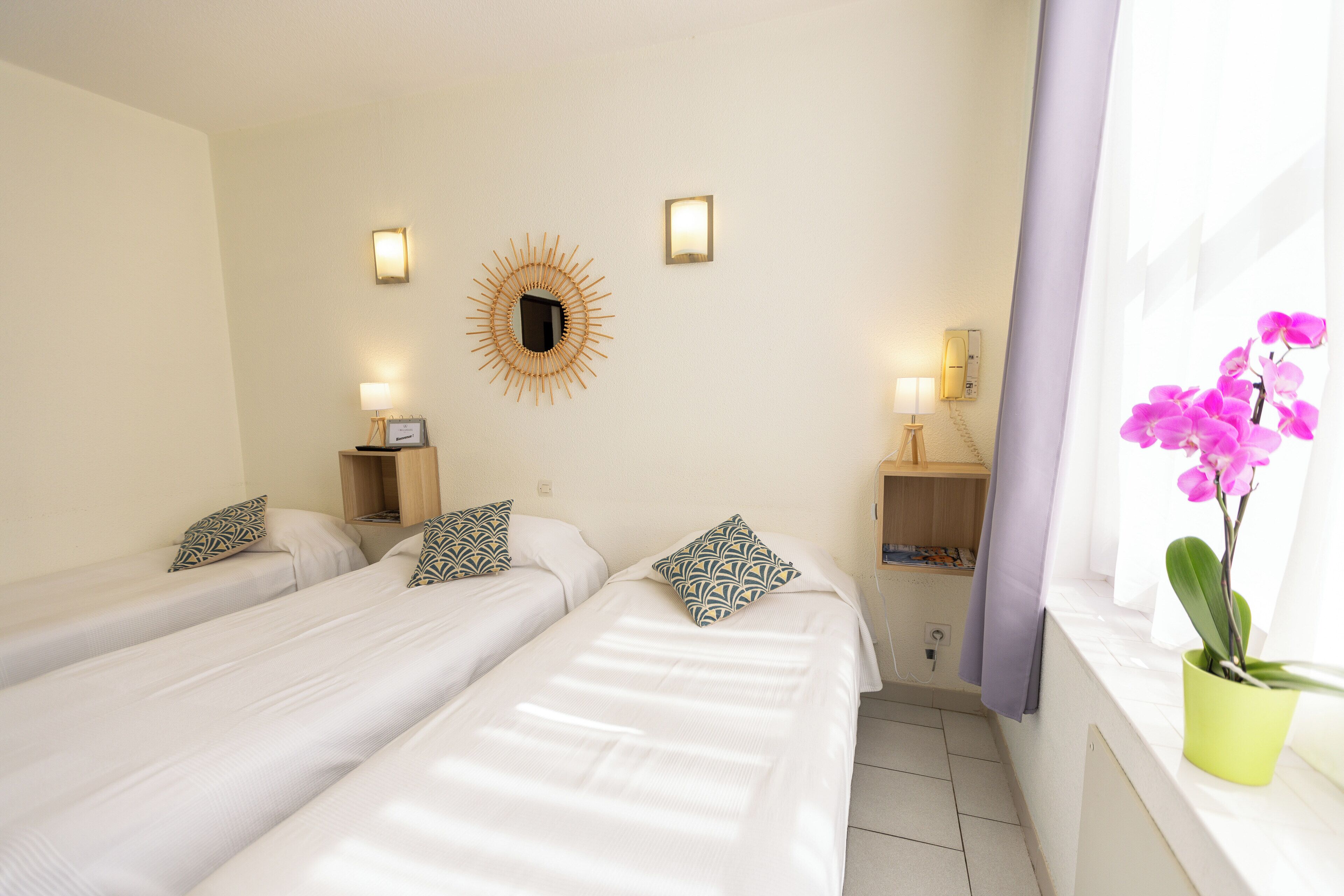 Quarto de hotel com duas camas de solteiro, decoração de parede em formato de sol, pequeno criado-mudo com abajur e janela com uma orquídea em vaso.
