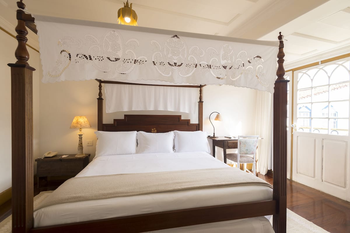 Una cama con dosel adornada con sábanas blancas, cabecera de madera y dosel de encaje. Hay mesitas de noche con lámparas y una silla en la habitación.