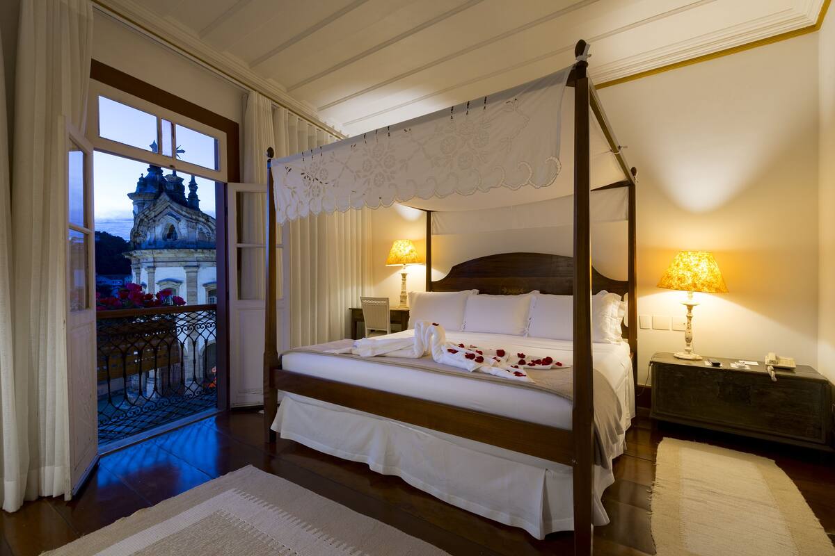 Una cama con dosel con sábanas blancas, dos lámparas de noche y vistas a un edificio histórico a través de un ventanal amplio.