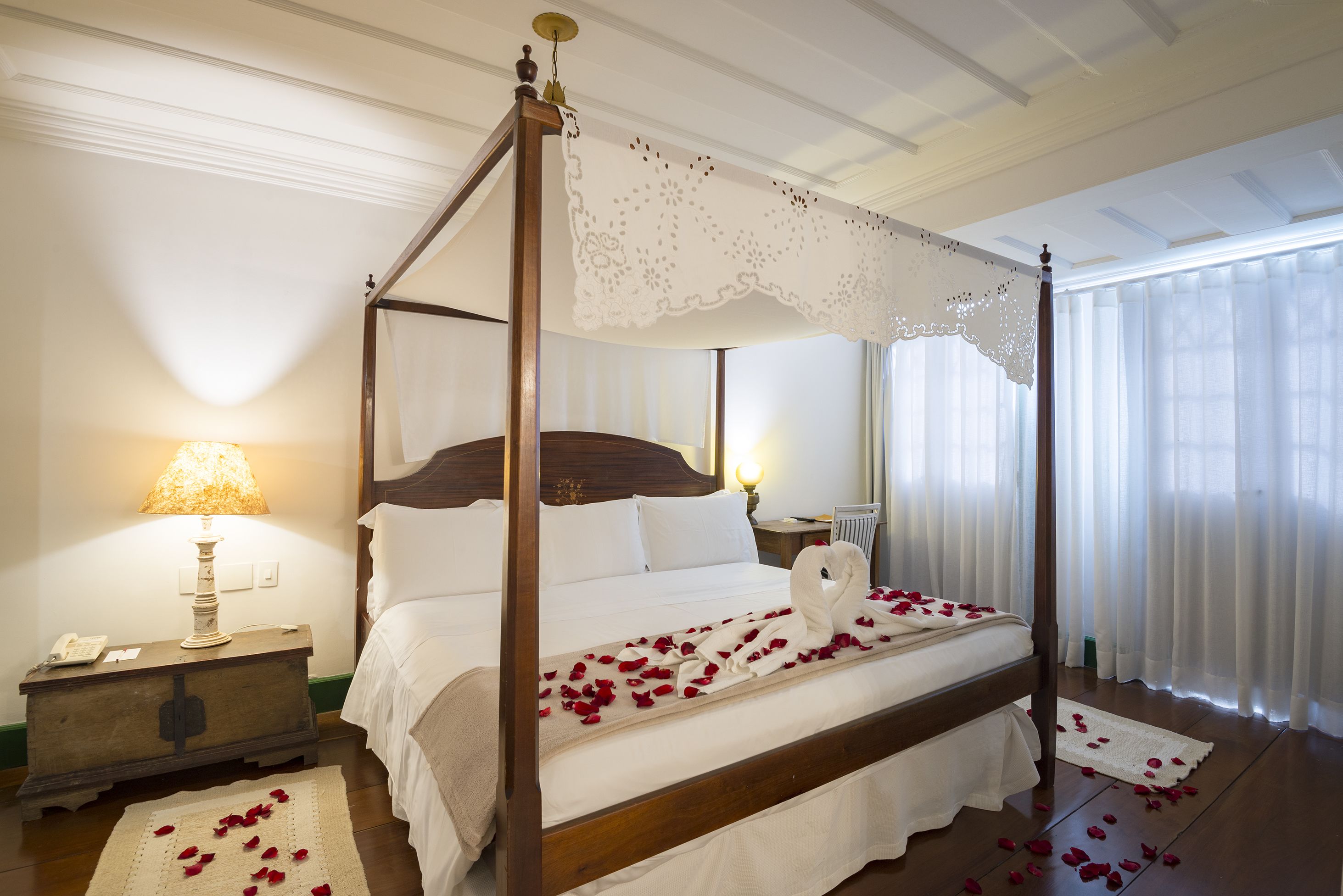 A four-poster bed with white linens and rose petals, a bedside lamp, and a wooden nightstand.