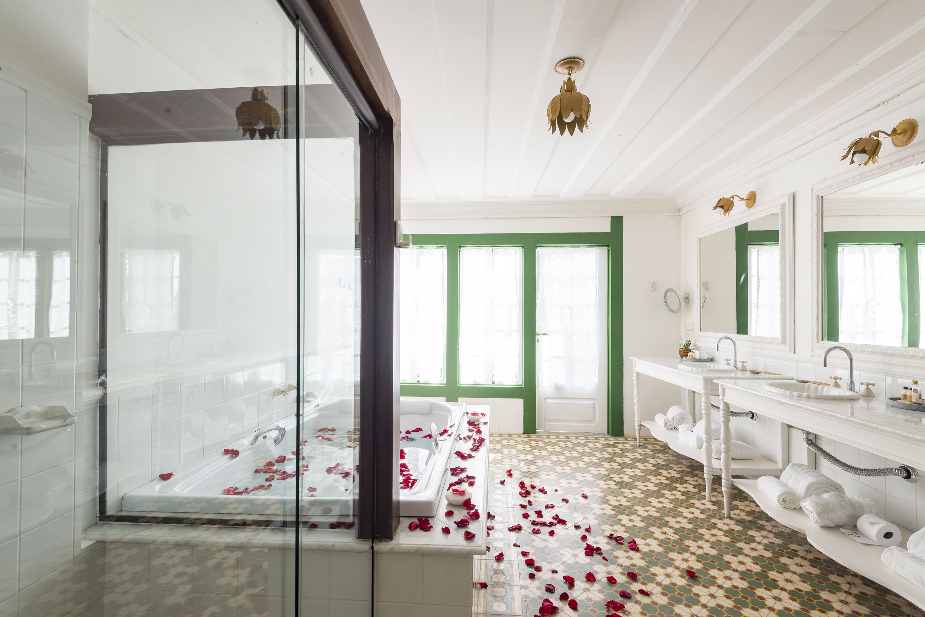 A bathroom with a glass shower enclosure, a bathtub with rose petals, a large mirror, and a patterned floor.