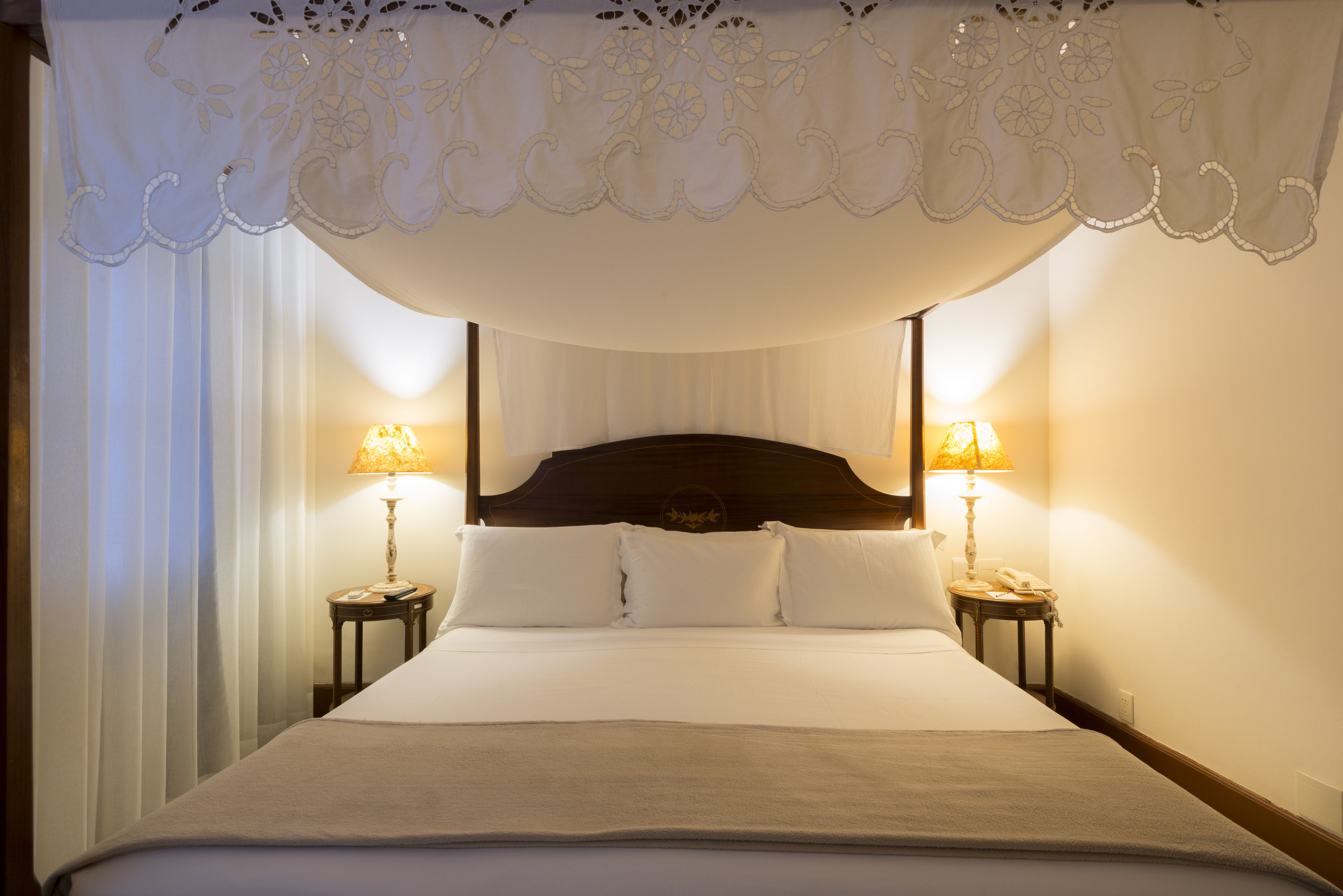 A neatly made bed with a headboard, two bedside tables with lamps, and sheer curtains.