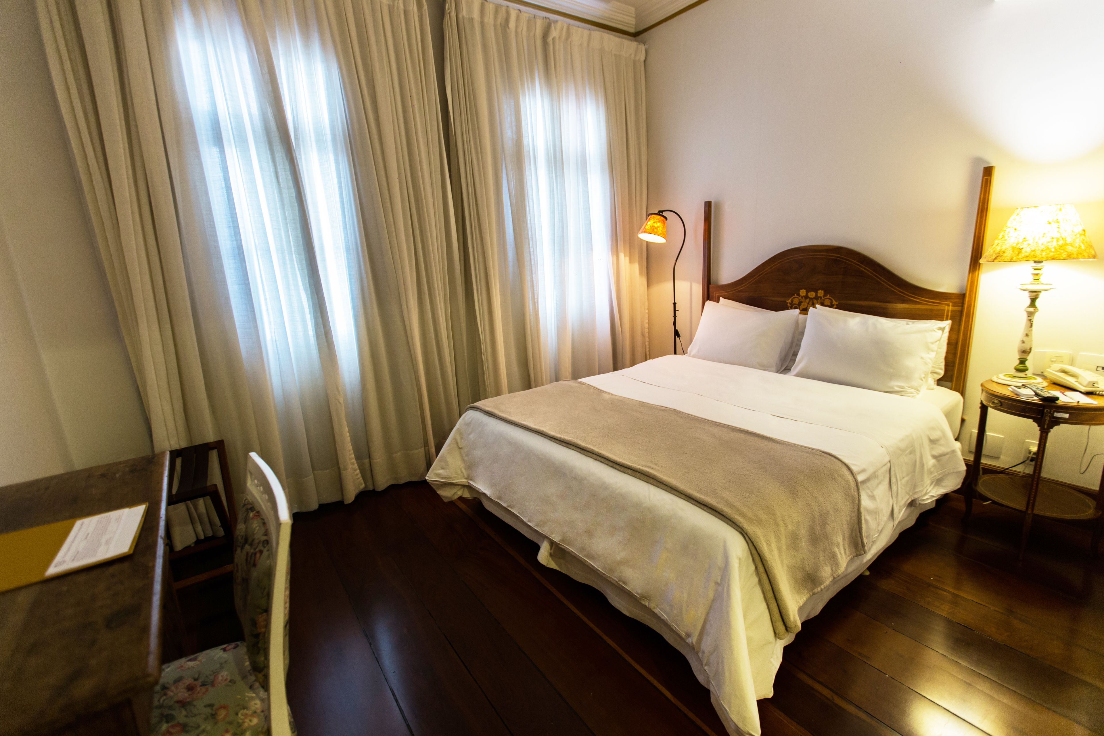 Quarto de hotel com cama, escrivaninha, cadeira, abajur e cortinas.