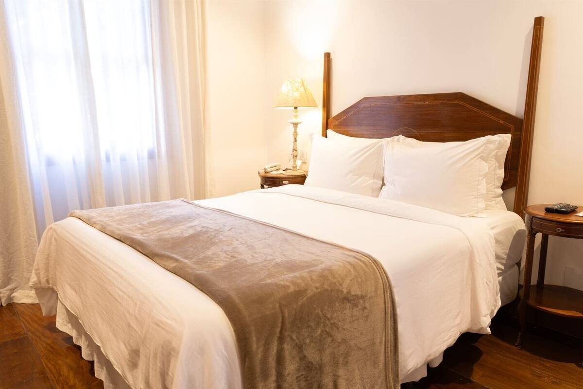 Una cama bien hecha con cabecera de madera, una mesita de noche con lámpara y una ventana con cortinas translúcidas.