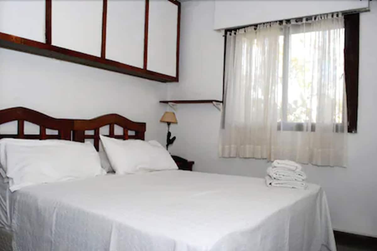 Una cama bien hecha con sábanas blancas, un cabecero de madera y una mesita de noche con una lámpara.