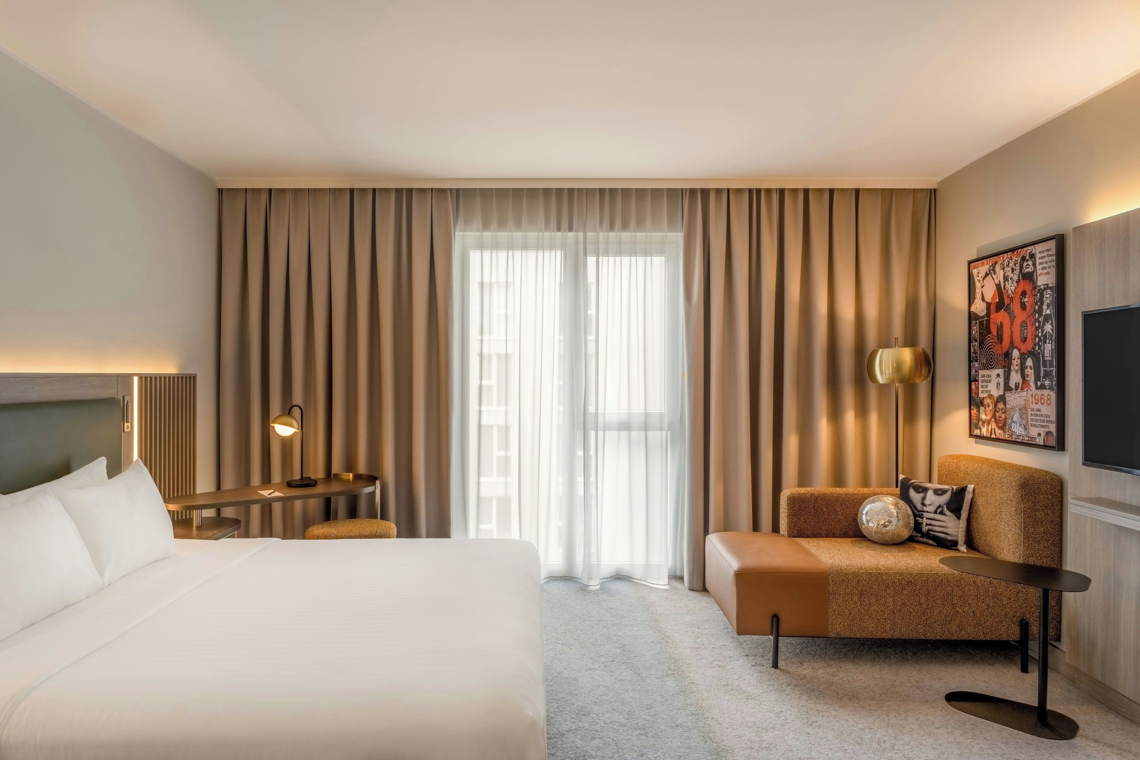 Ein Hotelzimmer mit einem großen Bett, einer Sitzecke mit Sofa und Sesseln, einem Schreibtisch mit Lampe und einem großen Fenster mit Vorhängen.