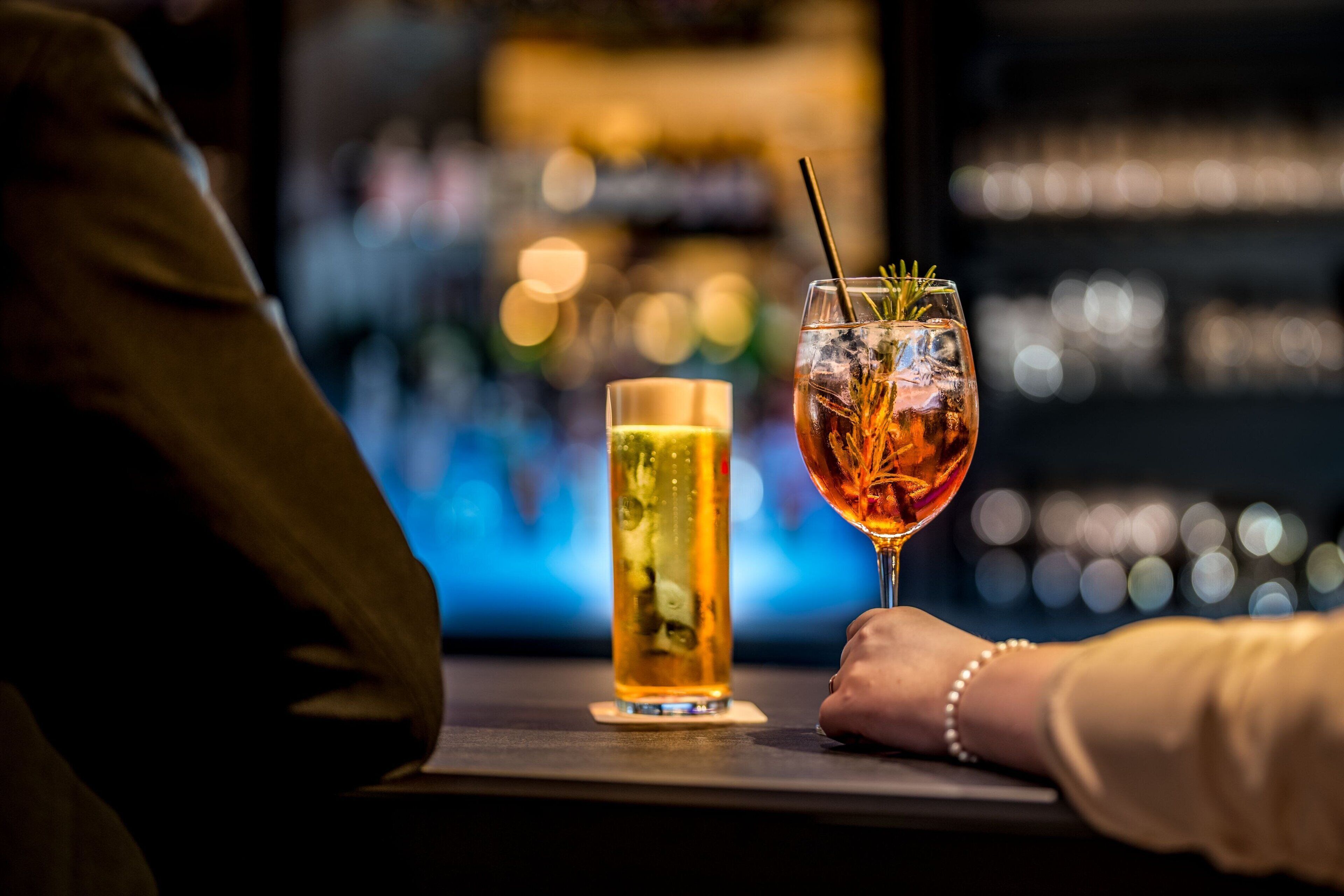 Un bar con dos bebidas sobre una mesa, una en un vaso alto y otra en uno más corto, con un fondo desenfocado que sugiere un ambiente animado.