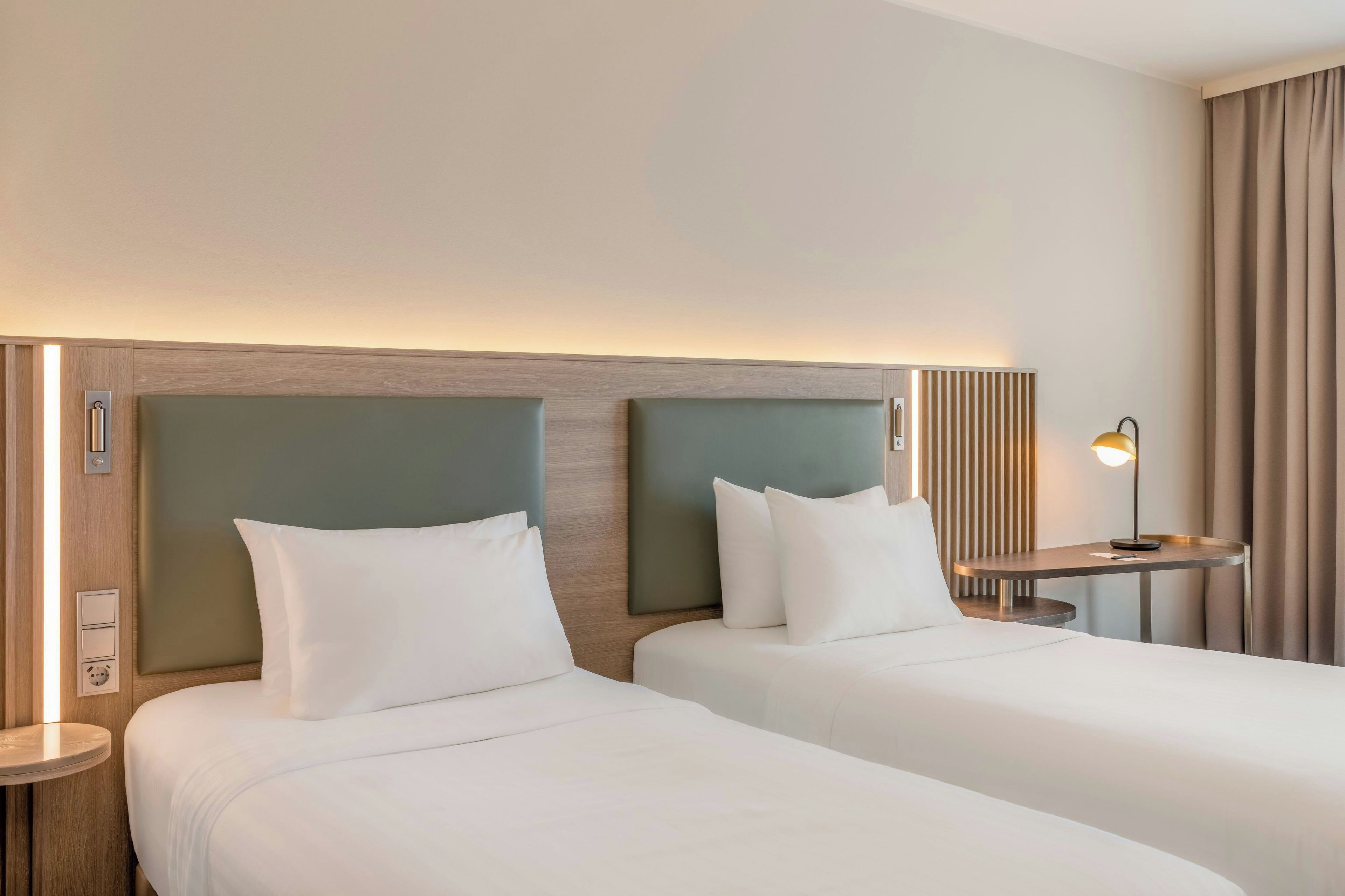 A hotel room with two beds, a bedside table with a lamp, and a wall-mounted light fixture.