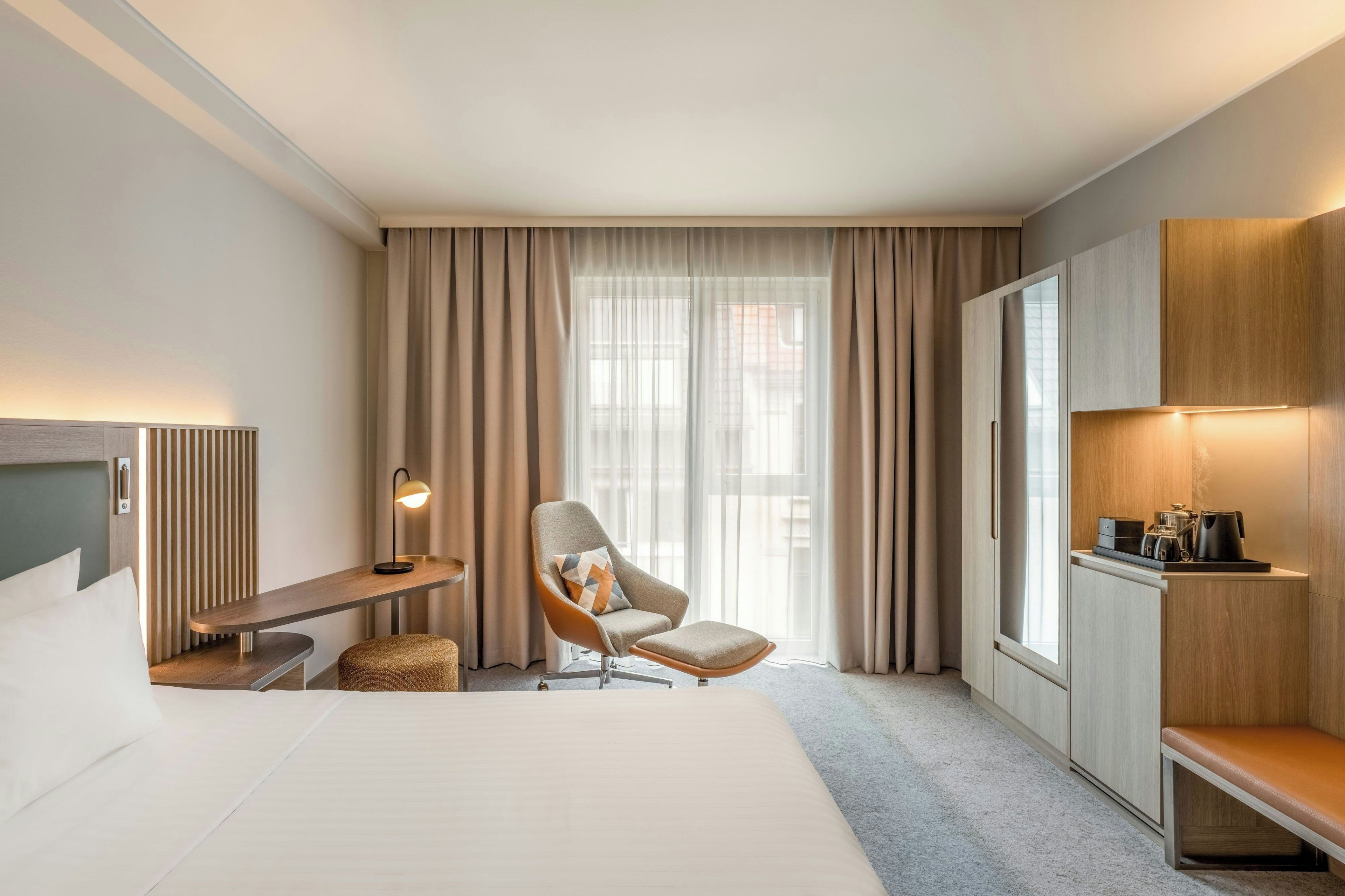 Habitación de hotel con cama, una silla, escritorio, televisor, ventana con cortinas y una zona de kitchenette.