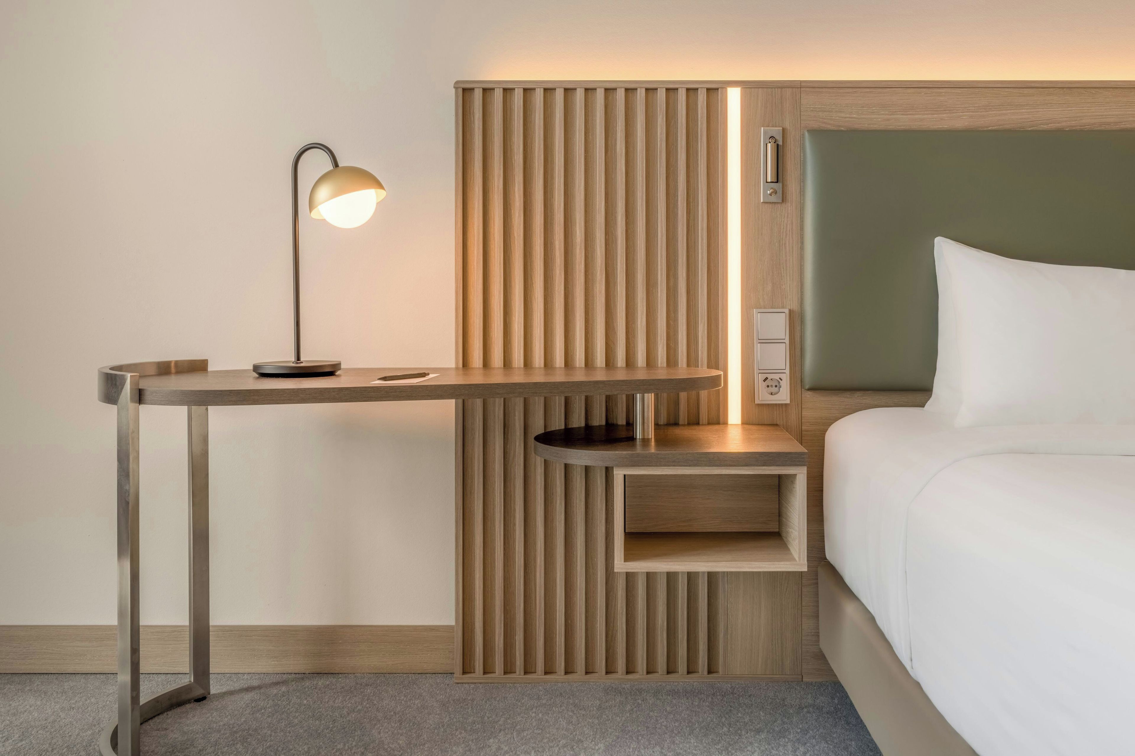 A modern hotel room with a wooden desk, a lamp, a bedside table, and a bed.