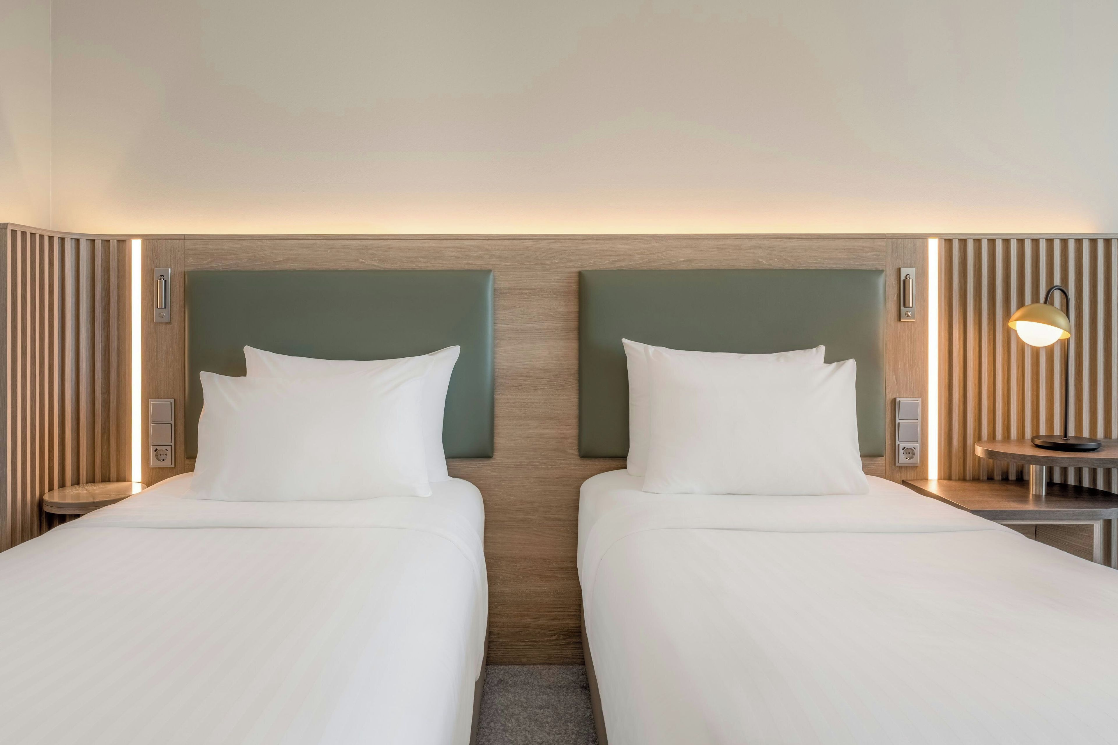 A hotel room with two single beds, a nightstand with a lamp, and a wall-mounted control panel.