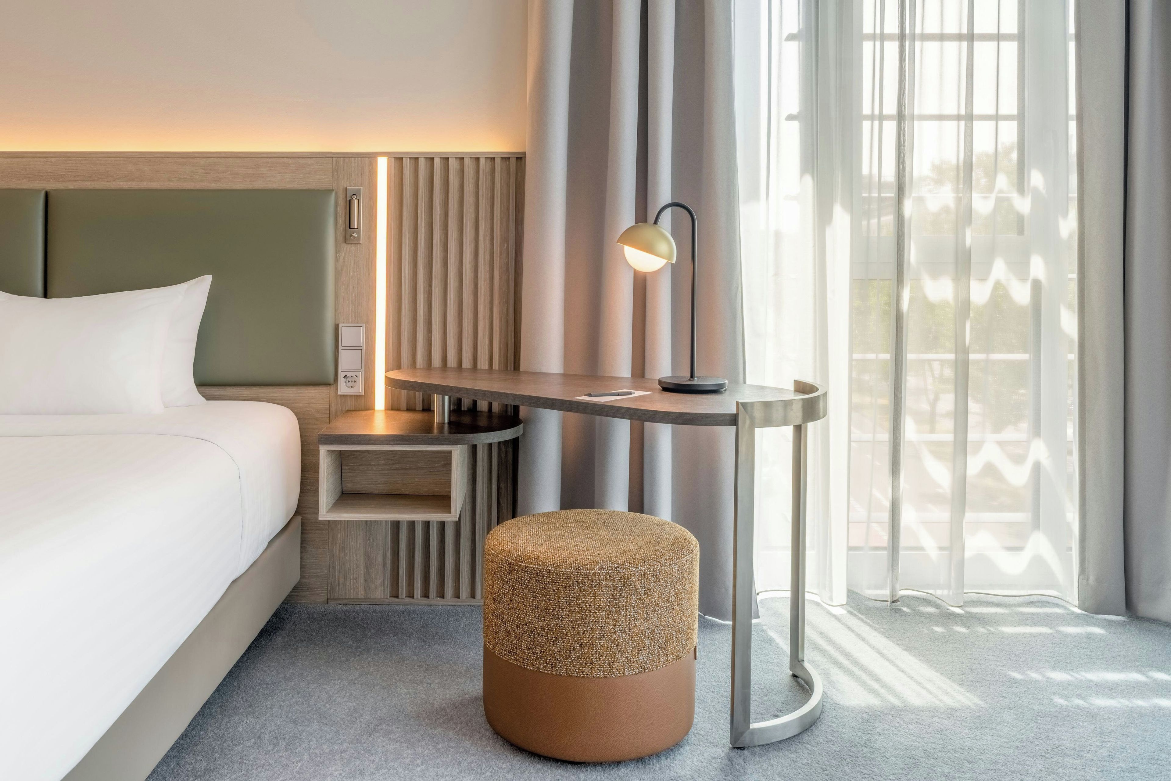 Habitación de hotel con cama, mesita de noche, lámpara y vistas al exterior a través de amplias ventanas.