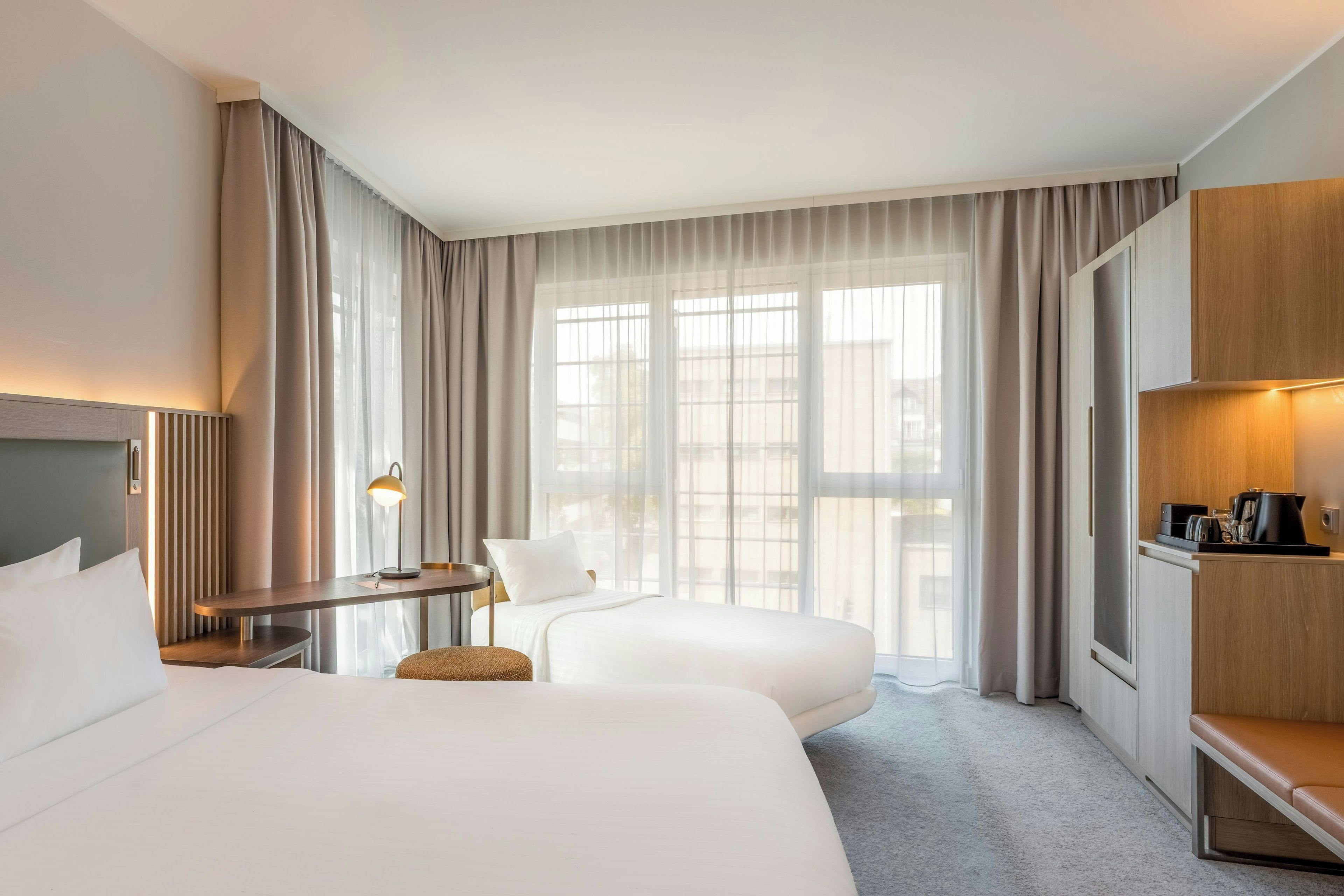 Habitación de hotel con dos camas, un escritorio, una silla, un pequeño reposapiés, cafetera y vistas de la ciudad a través de amplios ventanales.