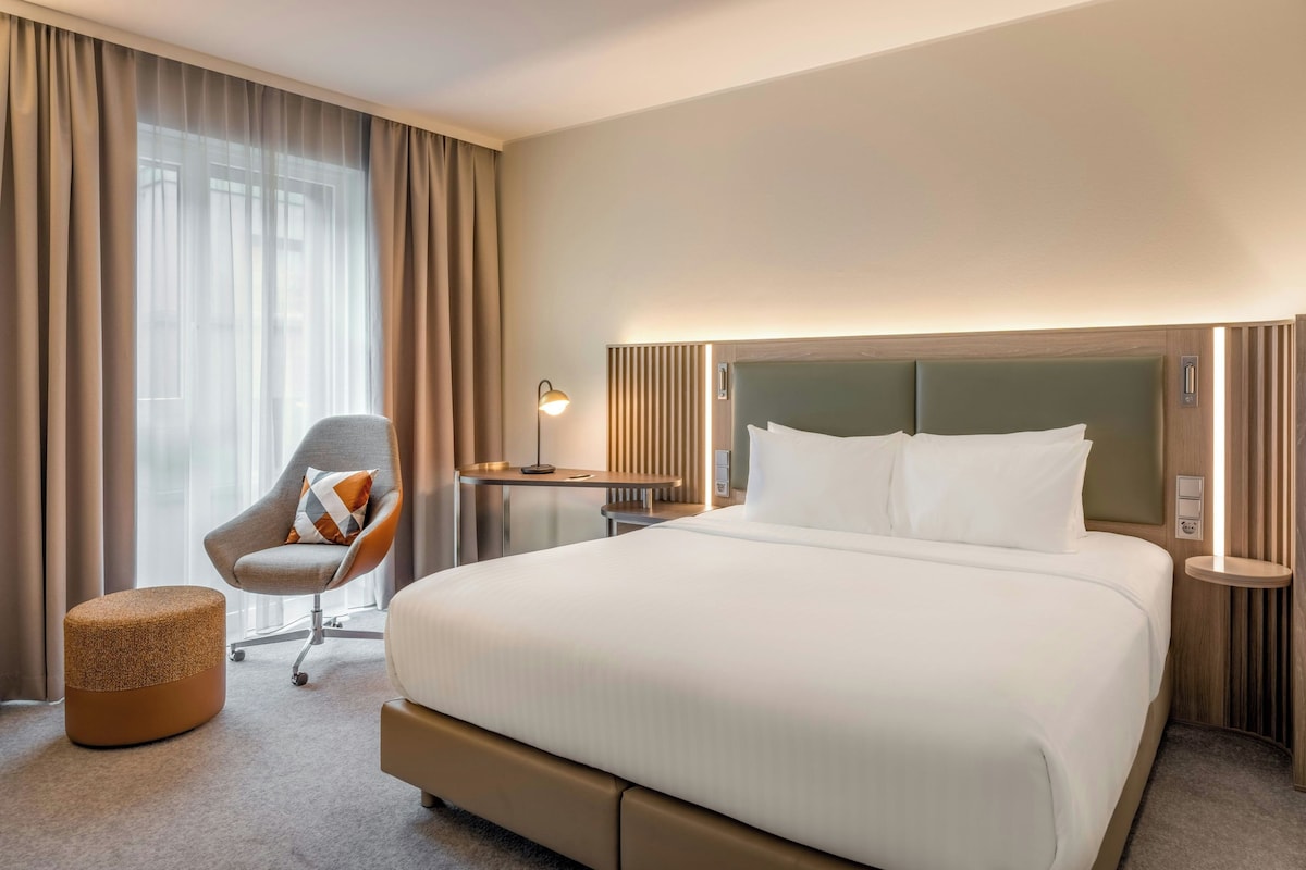 A hotel room with a large bed, a chair, a bedside table, and a lamp.