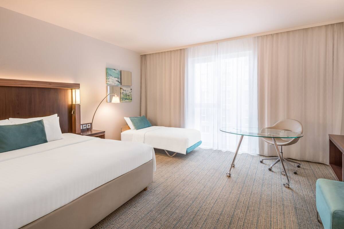 Une chambre d’hôtel avec un grand lit, un bureau avec une chaise, une petite table et une fenêtre avec des rideaux.