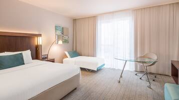 A hotel room with a large bed, a desk with a chair, a small table, and a window with curtains.