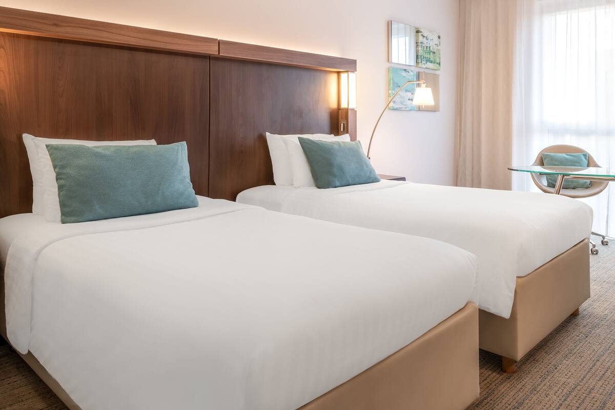 Une chambre d’hôtel avec deux lits, une tête de lit en bois, un bureau avec une chaise et une fenêtre avec des rideaux.