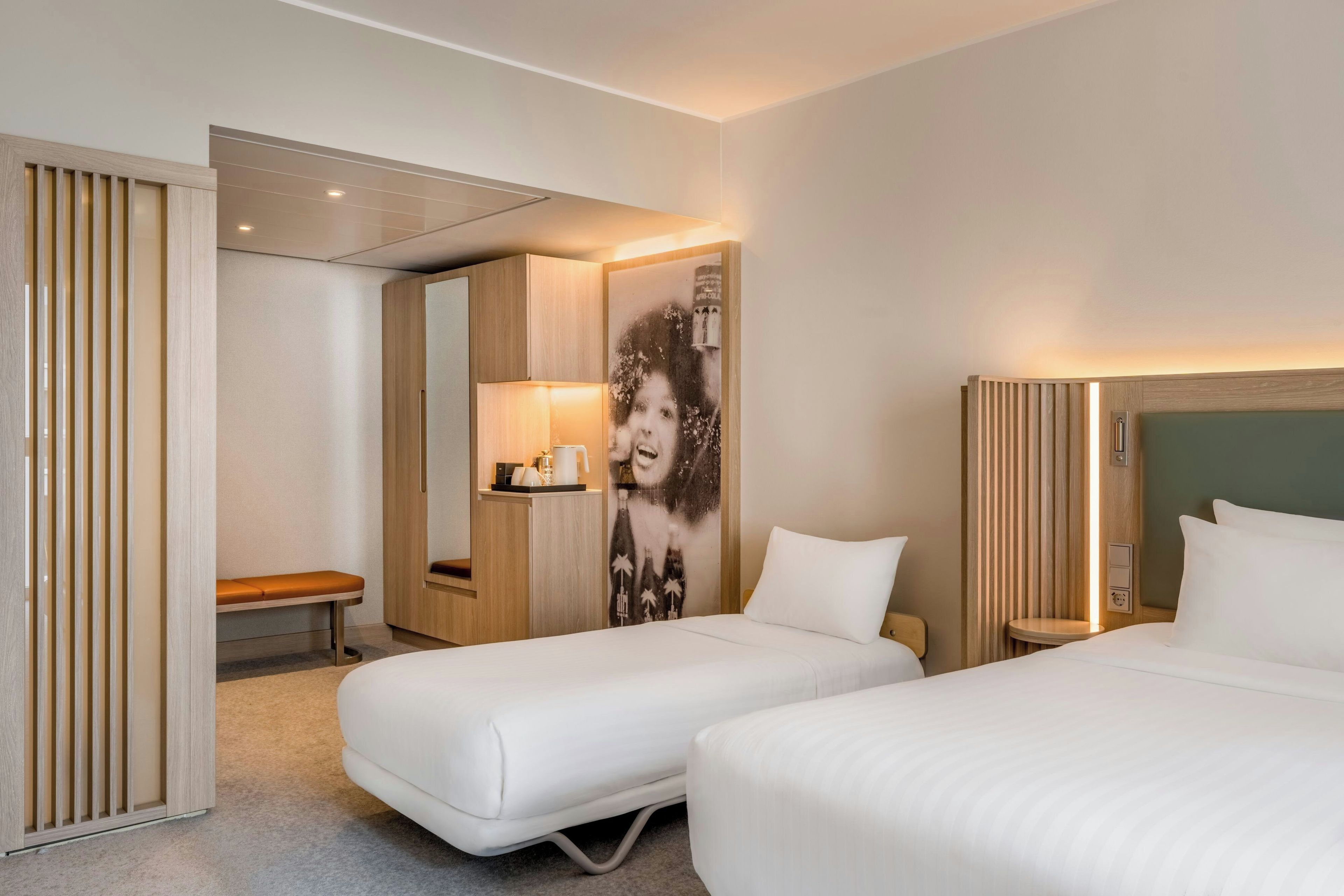 A hotel room with two beds, a bench, a wardrobe, and a wall with a large portrait.