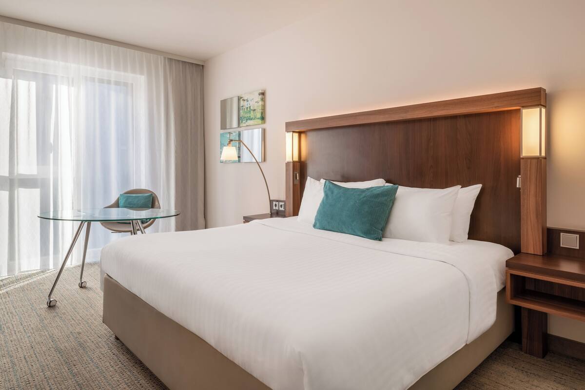 Une chambre d’hôtel avec un grand lit, une table de chevet, une petite table avec une chaise et une fenêtre avec des rideaux légers.