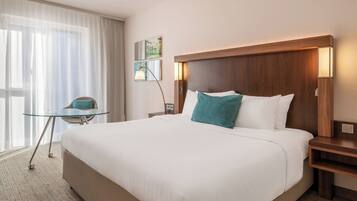 Habitación de hotel con una cama grande, una mesita de noche, una pequeña mesa con una silla y una ventana con cortinas traslúcidas.