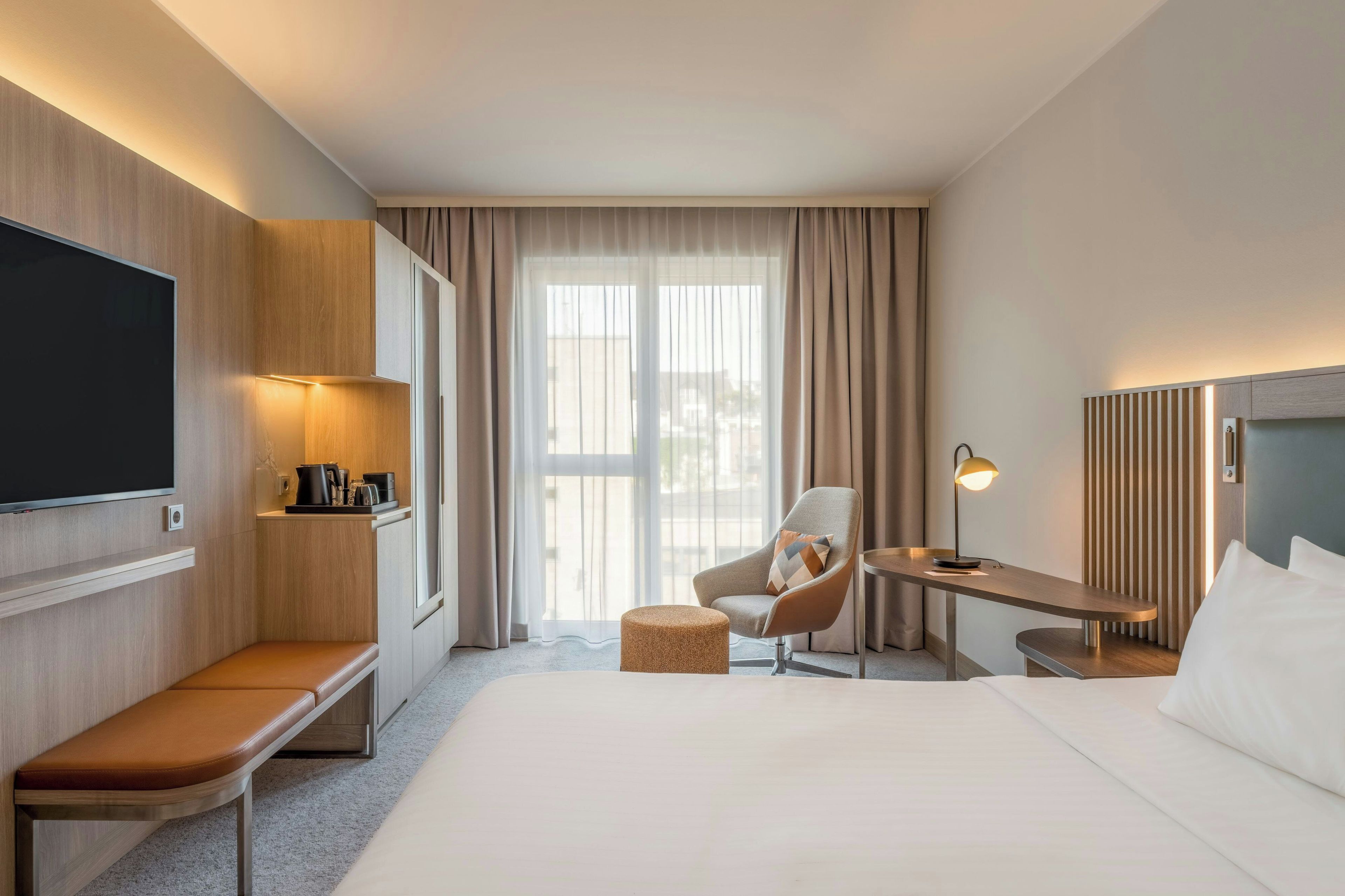 Una habitación de hotel con cama, escritorio, silla, televisor y ventana con cortinas.
