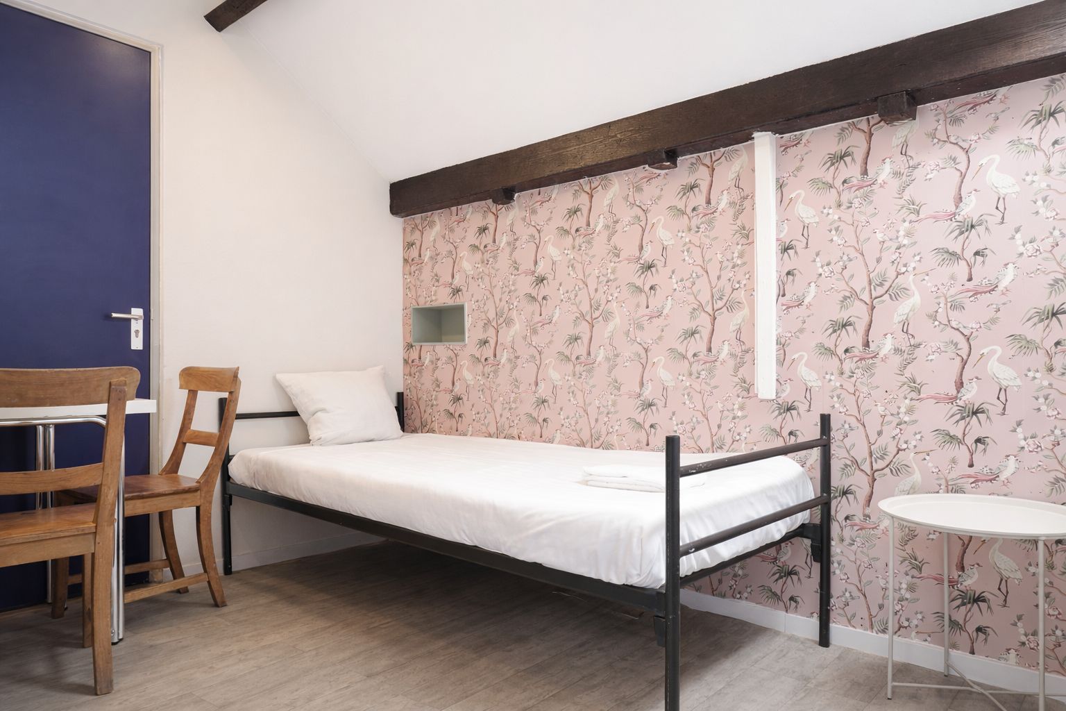 Un lit simple avec un cadre en bois, une petite table et une chaise dans une pièce dont la porte est bleue et dont un mur est recouvert d’un papier peint représentant des oiseaux tropicaux.