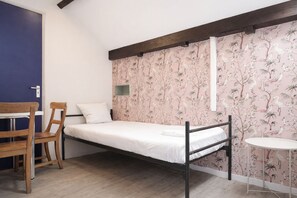 Un lit simple avec un cadre en bois, une petite table et une chaise dans une pièce dont la porte est bleue et dont un mur est recouvert d’un papier peint représentant des oiseaux tropicaux.