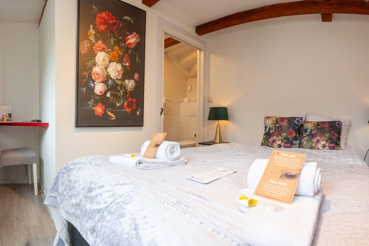 Habitación de hotel con cama, escritorio, silla, un cuadro y baño visible a través de una puerta abierta.