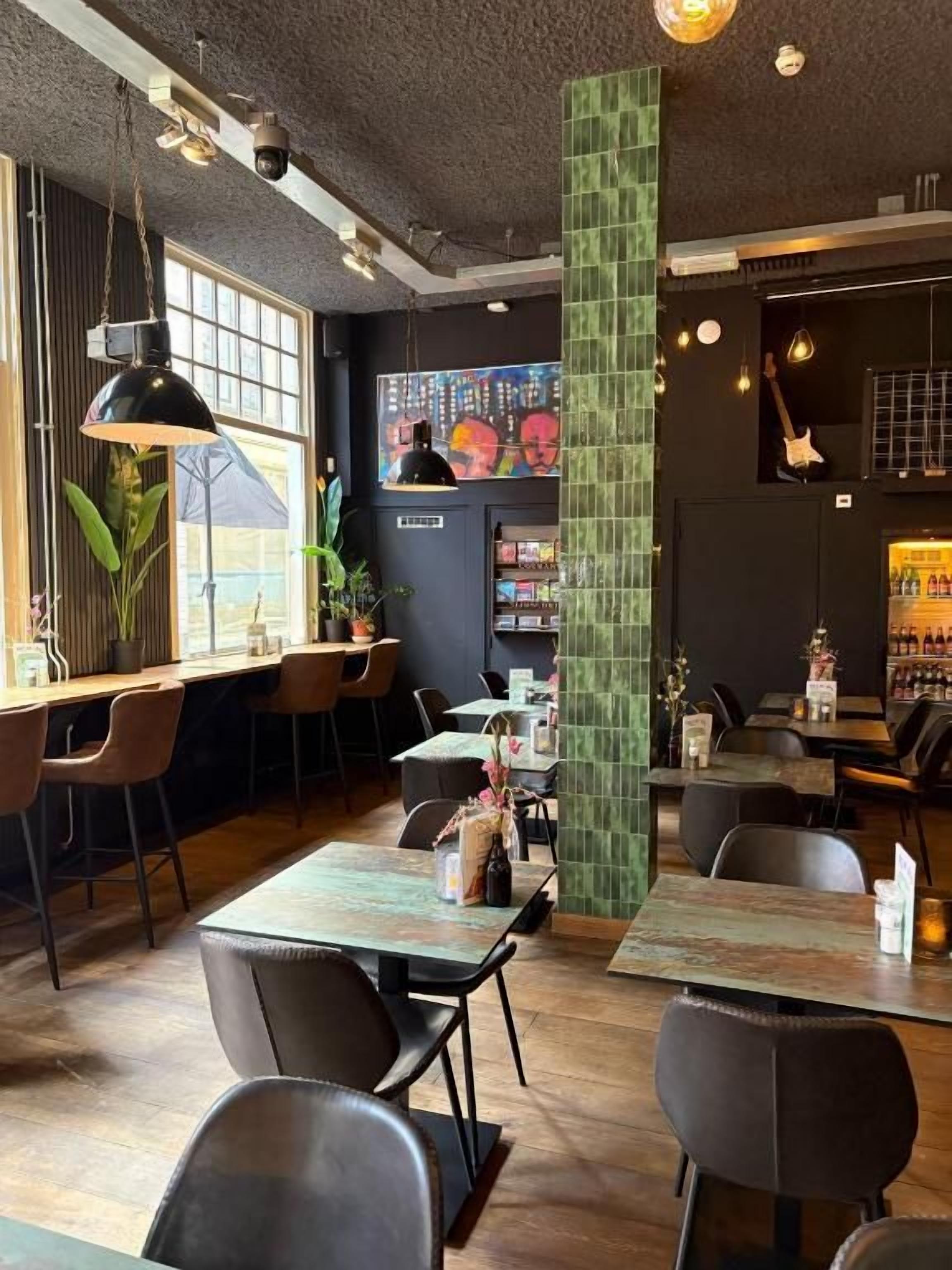 A restaurant interior with wooden tables, black chairs, hanging pendant lights, a green pillar, and a wall-mounted TV screen.
