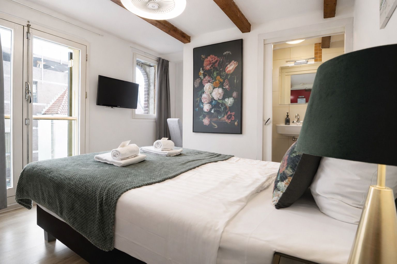 A bedroom with a bed, a flat-screen TV, a painting, a bathroom visible through an open door, and a window with curtains.
