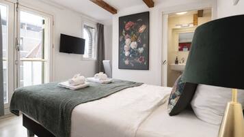 A bedroom with a bed, a flat-screen TV, a painting, a bathroom visible through an open door, and a window with curtains.