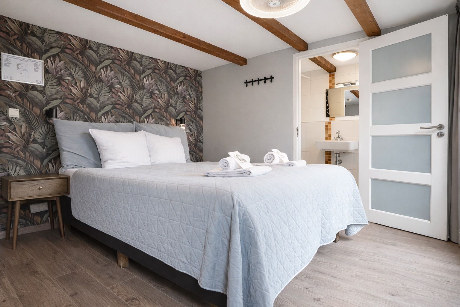 Een slaapkamer met een bed, een nachtkastje, een muur met bladmotief, een deur met geëtste glaspanelen, en een zichtbare badkamer door een open deur.