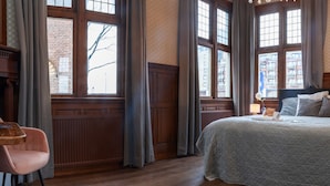 A hotel room with a large bed, a nightstand, a chair, and large windows with curtains.