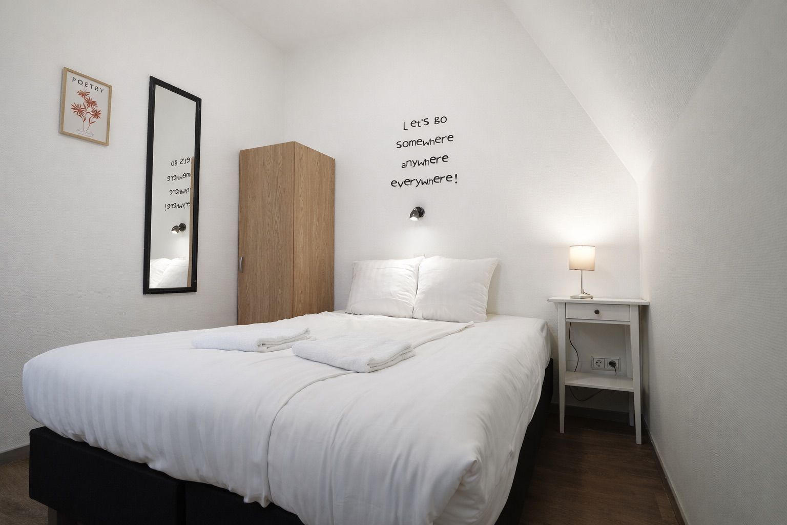 Een net opgemaakt bed met witte lakens, een houten garderobe, een nachtkastje met een lamp en een muur met een ingelijste quote.