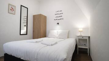 Een net opgemaakt bed met witte lakens, een houten garderobe, een nachtkastje met een lamp en een muur met een ingelijste quote.