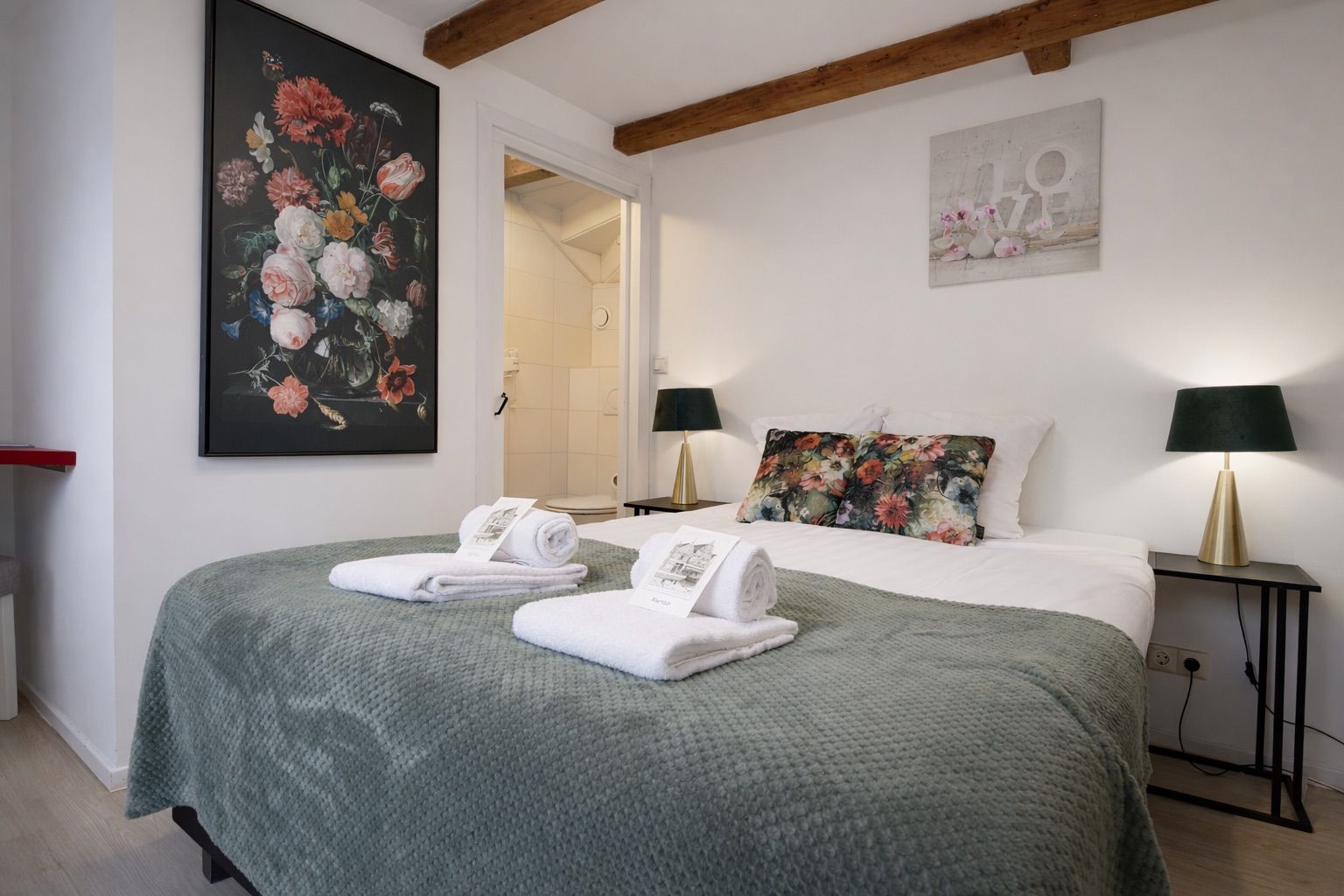 A hotel room with a bed, two bedside lamps, a painting, and a bathroom visible through an open door.