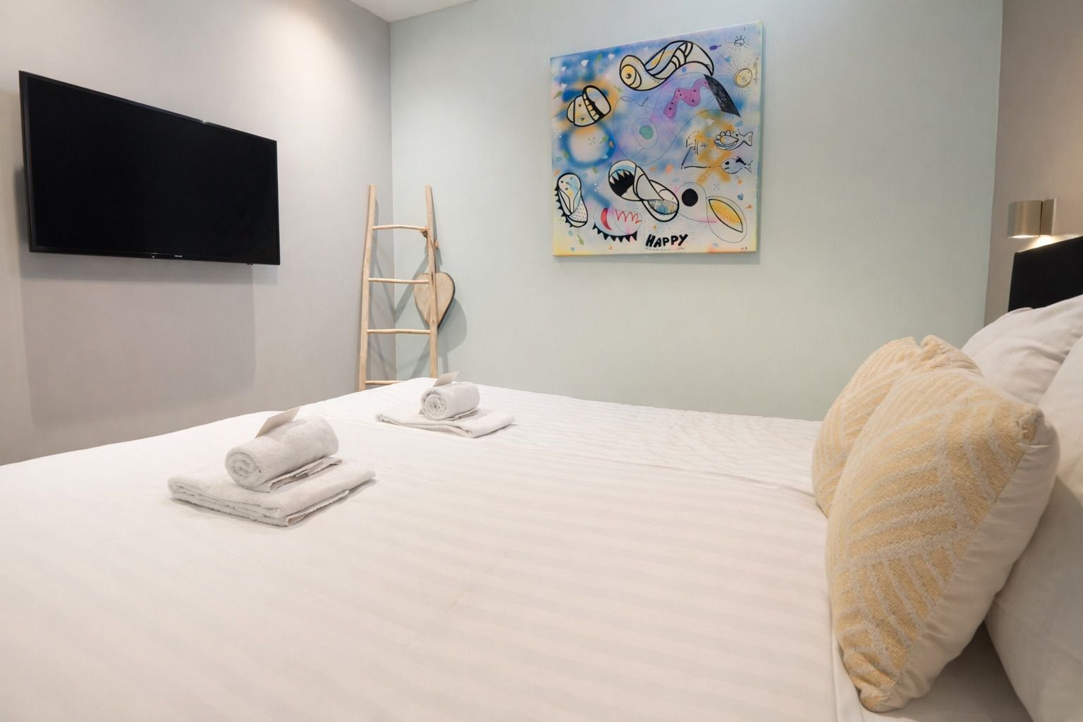 A hotel room with a bed, towels, a painting, a television, and a ladder.