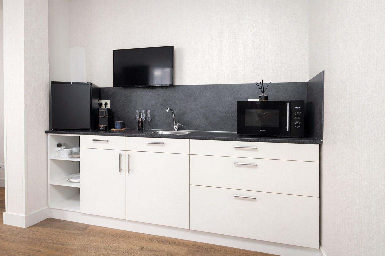 Een moderne keuken met witte kasten, een zwart aanrecht, een flatscreen televisie, een koffiezetapparaat en een magnetron.