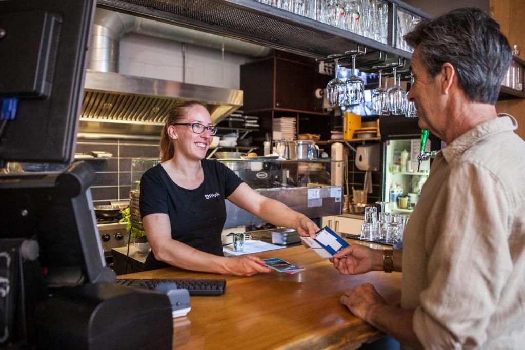 A bar setting with a bartender and a customer, a cash register, and a shelf with glasses.