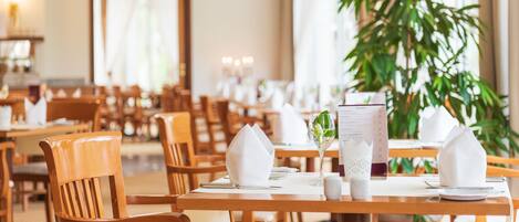Un restaurante con mesas y sillas de madera, vajilla blanca y una planta grande en maceta.