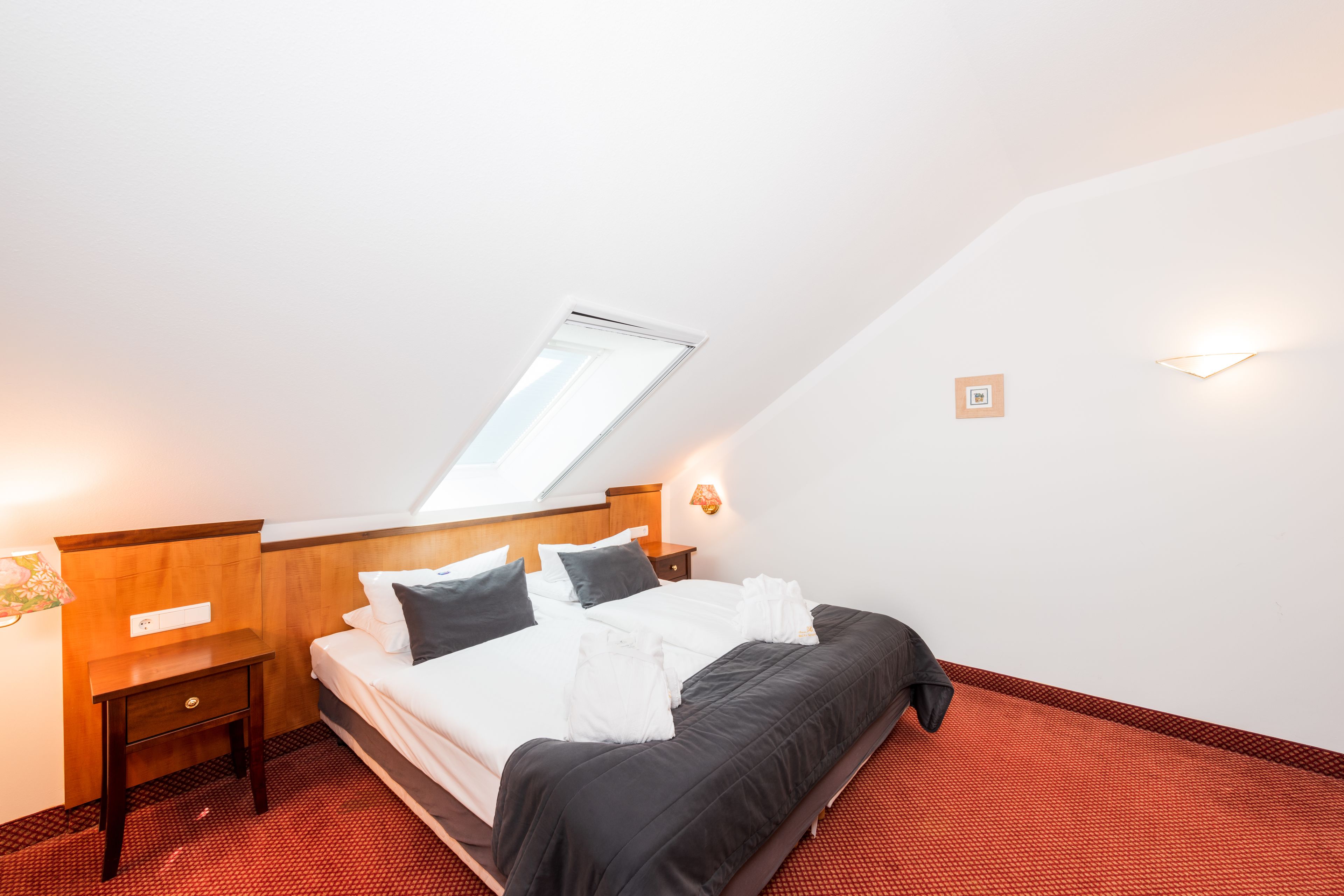 A neatly made bed in a room with a sloped ceiling, a small bedside table, and a wall-mounted light fixture.