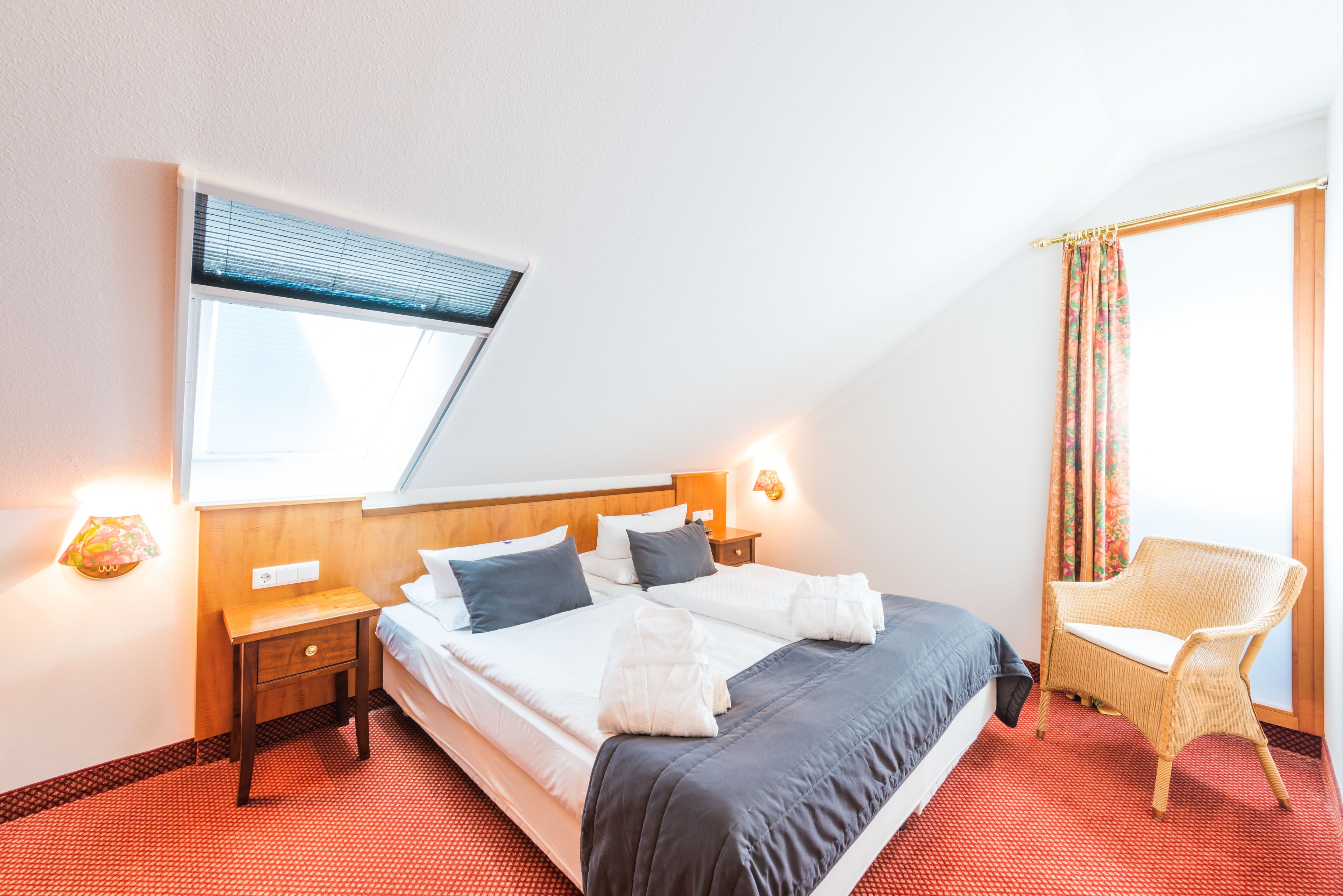 Habitación de hotel con una cama grande, una ventana con persianas, una silla y una mesita de noche con lámpara.